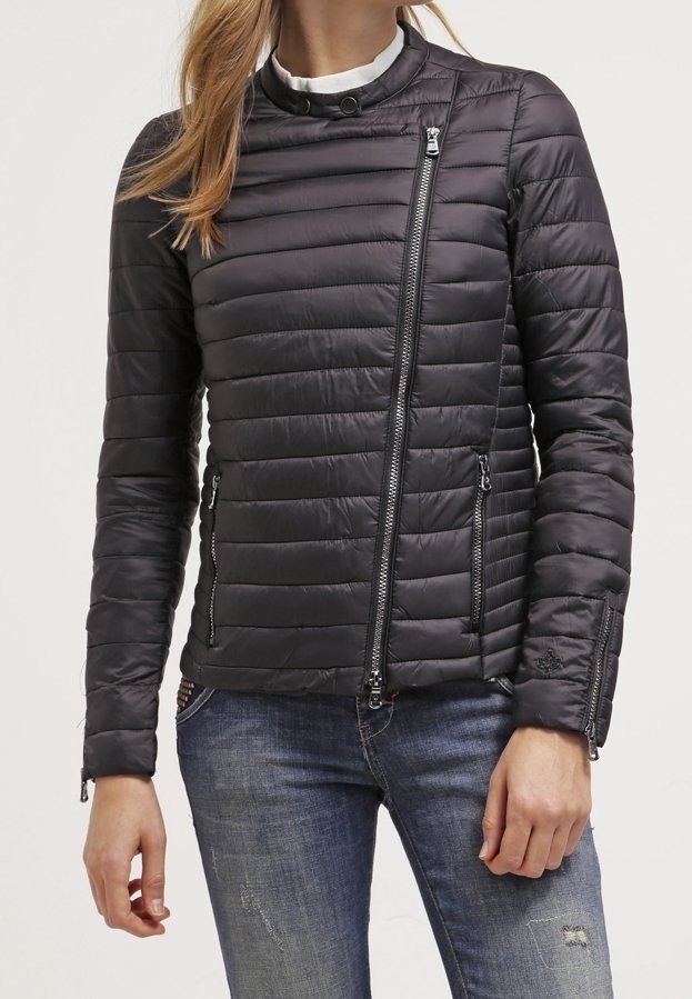 Femme portant une veste matelassée noire cintrée avec une fermeture éclair asymétrique sur le devant et des poches latérales zippées, associée à un jean bleu déchiré.