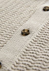 Cardigan en tricot beige avec des motifs texturés, deux boutons sombres et une couture verticale sur le devant. Matériau doux et chaud.