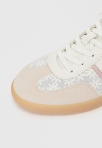 Chaussure avec un capuchon en suède beige, des lacets blancs, des panneaux latéraux transparents brodés de fleurs et une semelle en caoutchouc brun clair.