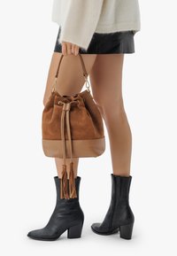 Brūns zamšs biroja soma ar ādas pamatni, sasienamu aizdari un pomponiem. Valkāta ar melnām augstpapēžu potīšu zābakiem un neitrālu džemperi.