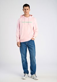 Rosa huva med "COPENHAGEN" i grön text, framficka, kombinerat med blå jeans och vita sneakers. Enkel, avslappnad design.