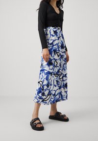 Černý top s dlouhým rukávem s knoflíkovými detaily, doplněný modrou a bílou vzorovanou midi sukní s květinovými motivy a předními knoflíky. Černé sandály.