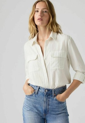 Mujer rubia con una camisa blanca de manga larga con bolsillos y vaqueros azules de talle alto, de pie con las manos en los bolsillos frente a un fondo liso.