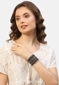 Brățară stratificată neagră și argintie cu textură împletită și accente metalice, purtată pe o mână împotriva unei bluze albe brodate.