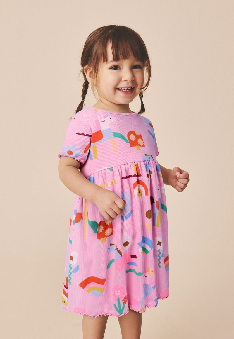Fille souriante aux cheveux bruns en couettes portant une robe rose avec des motifs colorés d’animaux, de fleurs et d’arcs-en-ciel sur un fond beige.