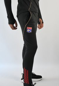 Pantalones deportivos negros con detalles en dorado, que presentan un parche de logo y franjas rojas a lo largo de los costados. Fabricados con un tejido elástico y texturizado.