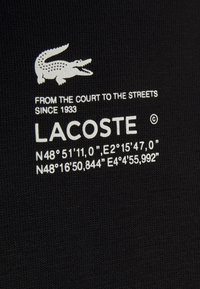 Material negru cu text alb, inclusiv "LACOSTE", coordonate și expresia "DE PE TEREN PE STRĂZI DIN 1933." Logo clasic prezent.