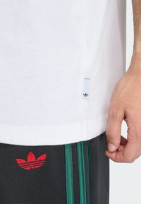 Λευκό βαμβακερό T-shirt με ετικέτα στην αριστερή πλευρά· κόκκινο κεντημένο λογότυπο στα γκρι παντελόνια με πράσινες ρίγες κατά μήκος των πλευρών.