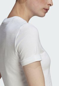 Balta medvilninė marškinė su apykakle ir suktomis rankovėmis, kuriomis išryškėja lygi tekstūra ir prigludęs siluetas.