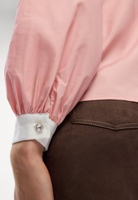 Blusa rosa chiaro con maniche a sbuffo e polsini bianchi con un bottone di cristallo rotondo. Tessuto marrone testurizzato visibile sottostante.