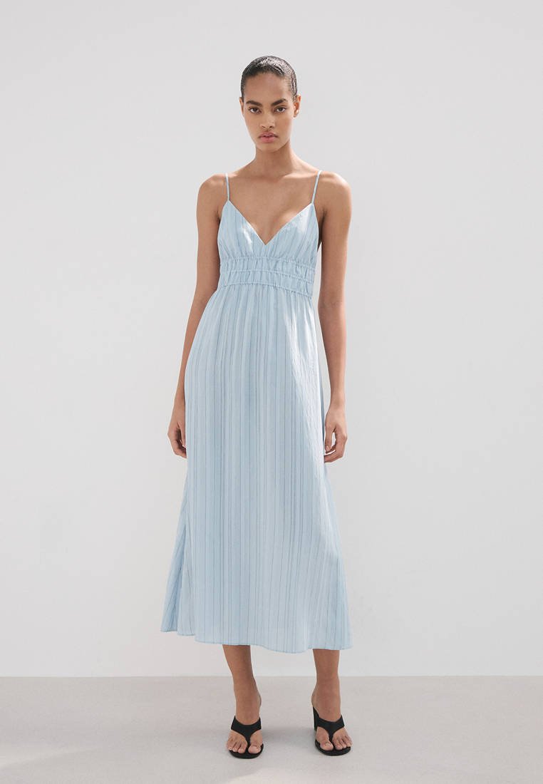 Femme portant une robe midi sans manches à rayures verticales bleu clair avec fines bretelles et talons ouverts noirs, debout devant un fond uni.