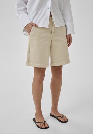 Žena oblečená v béžových krátkych nohaviciach po kolená, bielej košeli na gombíky a čiernych žabkách, stojaca s jednou rukou v vrecku.