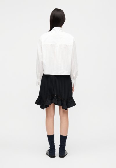 Άσπρο, μακρυμάνικο πουκάμισο με γιακά και υφή με λεπτές ρίγες, συνδυασμένο με μαύρη πλισέ φούστα με βολάν στο κάτω μέρος. Μαύρες κάλτσες και παπούτσια.