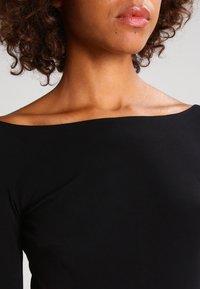 Haut noir à épaules dénudées, texture lisse, design ajusté, avec des manches longues et un large décolleté, mettant en valeur les épaules nues et la clavicule.