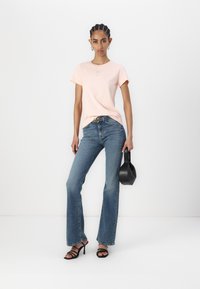 Gaiši rozā kokvilnas t-krekls ar īsām piedurknēm, kombinēts ar vidēji augstām zilo flared džinsu biksēm un melniem sandaliem ar siksniņām, tur rokā mazu melnu somiņu.