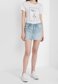 T-shirt bianco in cotone con un disegno minimalista di un volto in linea nera, abbinato a una mini gonna in denim blu chiaro e sneakers bianche.