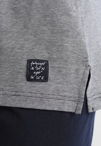 T-shirt rayé gris et blanc en coton, avec une fente sur le côté et une étiquette cousue portant des coordonnées et le nom de la marque.