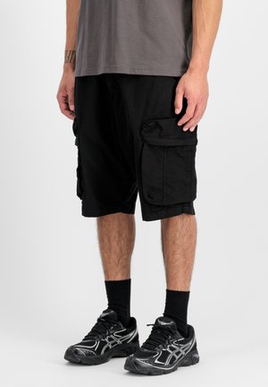 Homme portant un short cargo noir, des chaussettes noires et des chaussures de sport noires, debout devant un fond blanc uni.