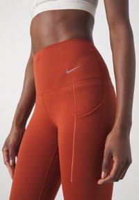 Atletyczna kobieta w brązowych legginsach Nike o wysokim stanie z boczną kieszenią oraz białym biustonoszu sportowym, stojąca na jednolitym tle.