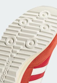 Punasilta punaiset Adidas-kenkien pohjat, joissa on valkoinen tekstuuripohja, jossa on pyöreitä pitoja ja Adidas-logo lähellä kantapäätä.