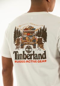 Camiseta blanca con un gráfico de un vehículo estacionado con equipo de camping, rodeado de árboles y montañas. El texto dice "Timberland RUGGED ACTIVE GEAR."