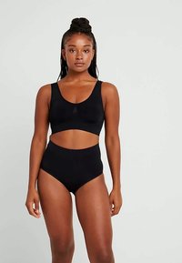 Kvinne med flettet hår som står, iført en svart ermeløs crop top og høyt liv undertøy foran en ensfarget lys bakgrunn.