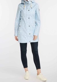 Femme portant un imperméable à capuche bleu clair à pois, un jean skinny noir et des baskets blanches, debout sur un fond blanc uni.