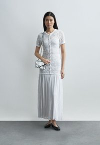 Žena stojí a má na sobě bílé krajkové šaty na knoflíky s plisovanou sukní, drží stříbrnou kabelku a nosí černé baleríny na šedé podlaze.