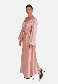 Lang, lyserød satinmorgenkåbe med brede ærmer, bundet i taljen; har en glat tekstur og elegant fald. Båret over en tank top og sorte hæle.