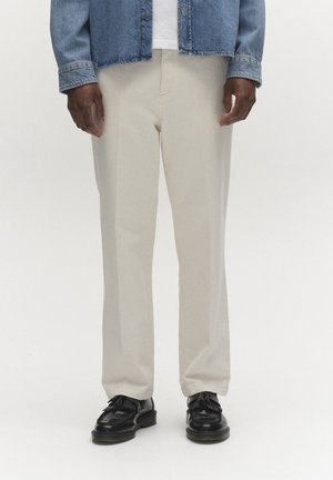 Mees kannab beežika sirge säärega pükse, musti tutiga mokasiine, valget särki ja sinist teksajope, taustal on lihtne valge taust.