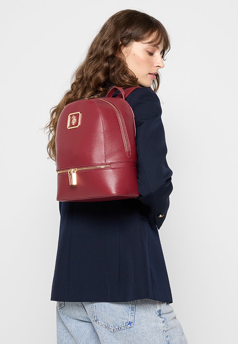 Zaino in finta pelle bordeaux con forma arrotondata, dettagli in metallo dorato e scomparto inferiore con zip, indossato su una spalla.