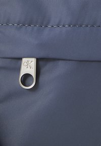 Ezüst cipzárhúzó "cK" logóval sötétkék texturált anyagon, közeli nézet egy táskával vagy ruhadarabbal.