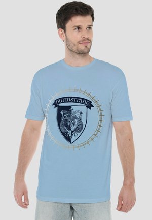 Man wearing light blue T-shirt with a Durmstrang crest featuring a horned animal skull and decorative circular pattern.