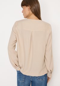 Beige langærmet bluse med rund halsudskæring, subtil bagplica og knapmanchetter, parret med blå denimjeans. Glat tekstur.