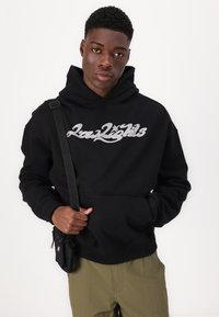Čierna mikina s kapucňou a trblietavým logom "Low Lights". Obsahuje klokaní vrecko, rebrované manžety a popruh cez rameno modela.