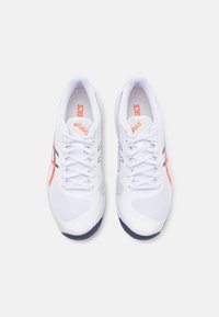 Zapatillas deportivas blancas con parte superior de malla, acentos en naranja y negro, punta redonda y suela oscura contrastante. Presenta un sistema de cordones tradicional.