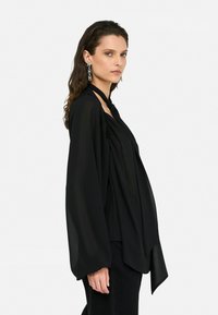 Blouse en chiffon noir avec de longues manches larges et un devant drapé. Dispose d'un lien au cou et de délicates fronces au niveau des épaules.