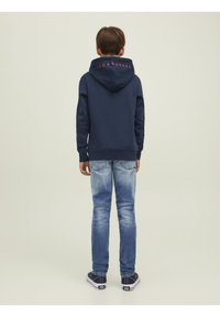 Námořnická mikina s velkou kapucí, na které je vzadu červený nápis "JACK & JONES". K ní jsou nošeny světle modré džíny a námořnické tenisky.