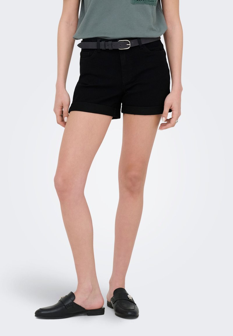 Femme portant un short noir à ourlet roulotté avec une ceinture noire, un haut vert et des mocassins noirs slip-on, debout devant un fond uni.