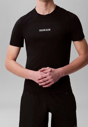 Mann trägt ein schwarzes Calvin Klein Kurzarm-T-Shirt und passende schwarze Hose, Hände in der Taille verschränkt, vor grauem Hintergrund.