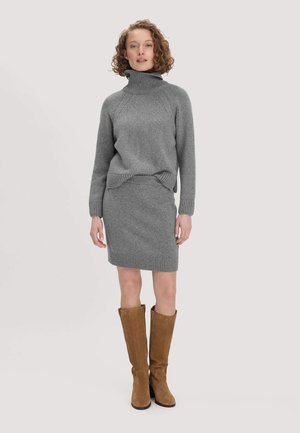 Frau mit lockigem Haar, die einen grauen Rollkragenpullover und einen Rock sowie braune kniehohe Stiefel trägt, steht vor einem schlichten, hellen Hintergrund.
