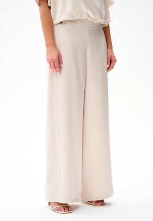 Kobieta w kremowych, szerokich spodniach z wysokim stanem, w zestawie z dopasowanym topem i beżowymi, otwartymi na palcach szpilkami, stojąca na tle białego tła.