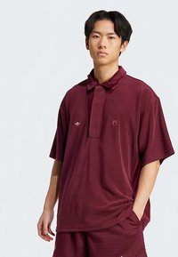 Mladý muž v voľnom vínovom polo tričku s krátkym rukávom a zodpovedajúcich šortkách, stojí s jednou rukou v vrecku na pozadí bez vzoru.