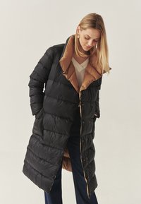 Giacca a vento nera con interno marrone, caratterizzata da un colletto alto, texture trapuntata e chiusure con bottoni a scatto. Design lungo e leggermente oversize.