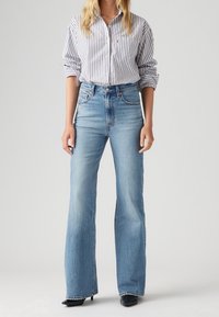 Jeans de tiro alto azul claro con pierna acampanada, con un dobladillo sin acabar. Combinados con una camisa de botones a rayas blanca y azul marino.