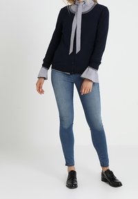 Žena v námořnickém cardiganovém svetru přes pruhovanou blůzu s kravatou, doplněná modrými skinny džíny a černými šněrovacími botami.