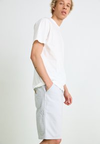 Lichtgrijze shorts met een relaxed fit, zijzakken en een patchlogo. Combinatie met een eenvoudig wit katoenen T-shirt. Eenvoudige achtergrond.