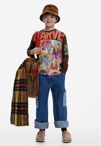 Băiat îmbrăcat într-un tricou maro cu mâneci lungi și personaje de benzi desenate colorate, pantaloni din denim cu manșete, pălărie maro din catifea tip bucket și o geantă roșie.