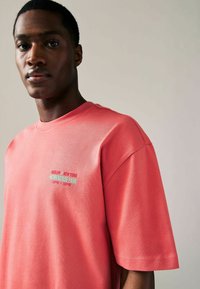 Koralfarvet t-shirt lavet af blødt stof, med rund halsudskæring og løs pasform. Fremtrædende tekstgrafik i turkis på brystområdet.