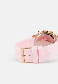Bracelet de montre en caoutchouc rose avec une boucle en ton or et un boîtier en or rose, présentant un cadran rond et des détails texturés sur le bracelet.